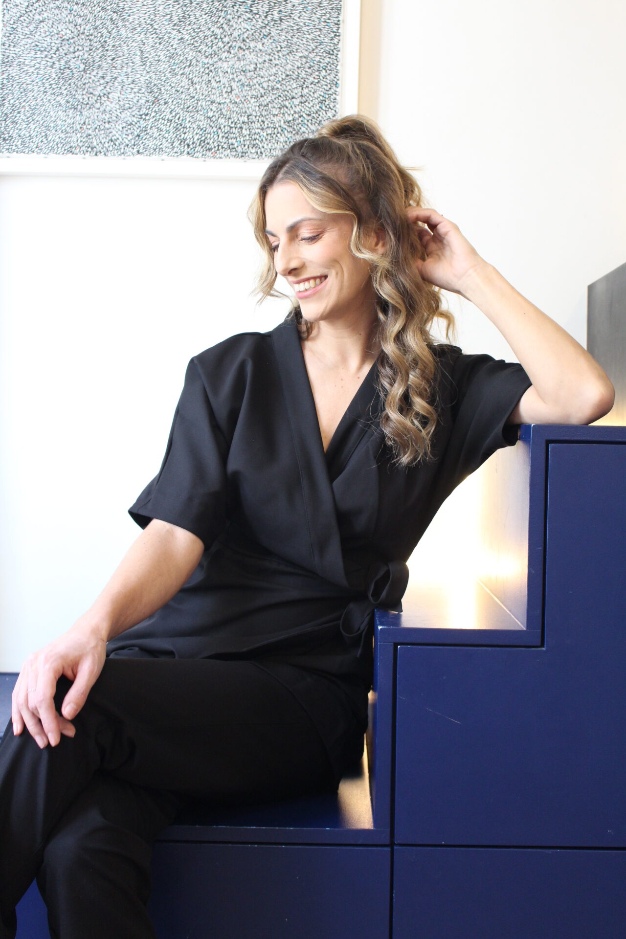 Femme souriante assise sur des marches bleues, portant une blouse noire élégante et dégageant une coiffure légère. Femme souriante assise sur des marches bleues, portant une blouse noire élégante et dégageant une coiffure légère.
