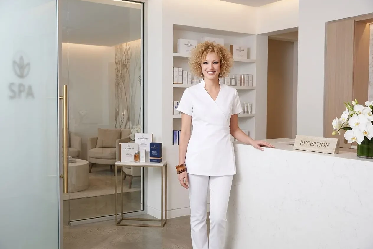 tenue professionnelle esthéticienne institut beauté: femme en blouse blanche et pantalon blanc devant une réception d’institut spa, ambiance lumineuse et accueillante